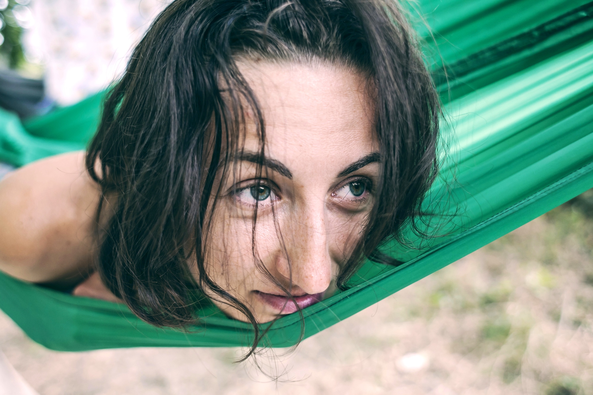 A person with wavy brown hair is resting in a green hammock, gazing thoughtfully into the distance. The background is blurred, suggesting an outdoor setting with greenery.