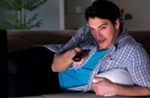 A man lounges on a sofa, holding a remote control and looking at the TV. He is wearing a plaid shirt over a blue t-shirt and lying on his side with a white pillow under his arm. A plant is visible in the background.