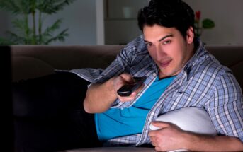 A man lounges on a sofa, holding a remote control and looking at the TV. He is wearing a plaid shirt over a blue t-shirt and lying on his side with a white pillow under his arm. A plant is visible in the background.