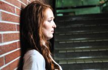 A woman with long red hair leans against a brick wall, looking contemplative. She is wearing a white shirt, and the background shows a staircase with dim lighting.