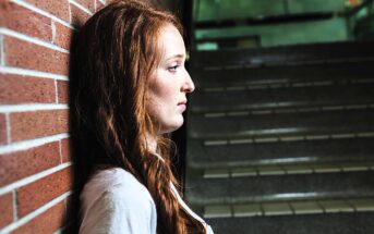 A woman with long red hair leans against a brick wall, looking contemplative. She is wearing a white shirt, and the background shows a staircase with dim lighting.