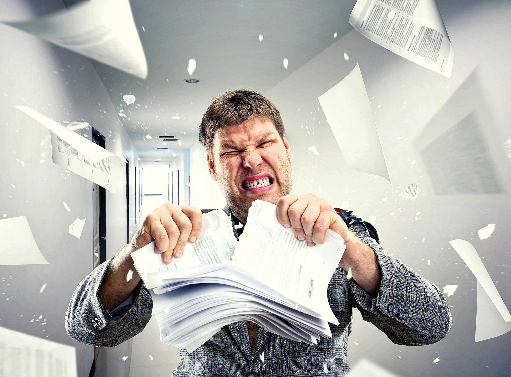 A man in a suit angrily tearing apart a stack of papers in a hallway. Papers are flying around him, creating a chaotic scene. His facial expression shows frustration and intensity. The hallway is brightly lit.