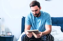 A man with a beard sits on a bed, wearing a blue t-shirt and striped pants, looking thoughtfully at a framed photo he's holding. The room is softly lit, with a white wall and a navy headboard in the background.