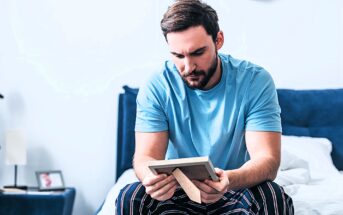 A man with a beard sits on a bed, wearing a blue t-shirt and striped pants, looking thoughtfully at a framed photo he's holding. The room is softly lit, with a white wall and a navy headboard in the background.