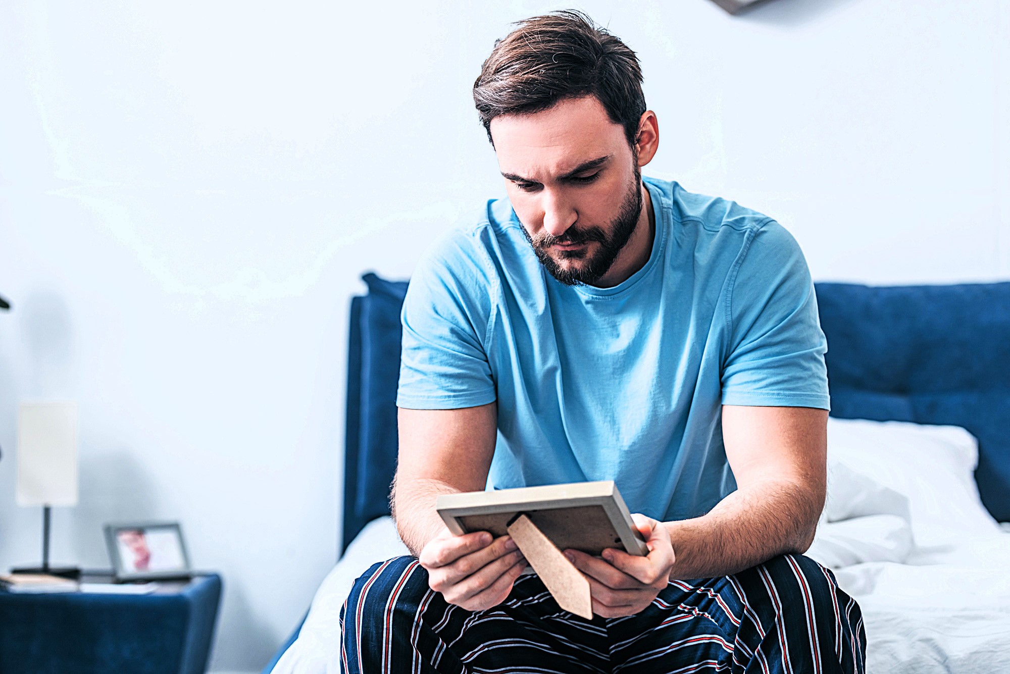 A man with a beard sits on a bed, wearing a blue t-shirt and striped pants, looking thoughtfully at a framed photo he's holding. The room is softly lit, with a white wall and a navy headboard in the background.