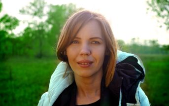 A woman with shoulder-length brown hair stands outdoors in a green field, wearing a light-colored jacket. The sun is shining from behind, creating a soft glow around her head.