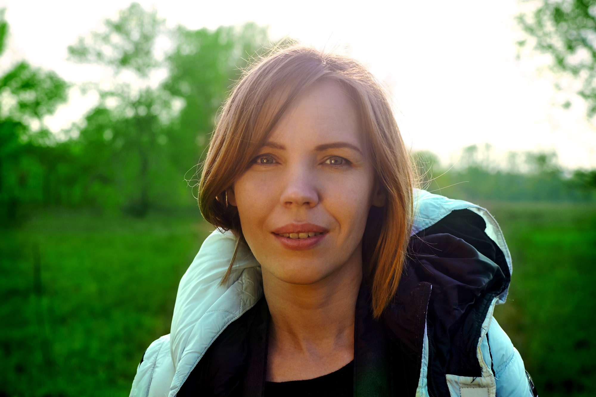 A woman with shoulder-length brown hair stands outdoors in a green field, wearing a light-colored jacket. The sun is shining from behind, creating a soft glow around her head.