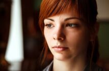 A young woman with red hair and a contemplative expression looks slightly away from the camera, set against a softly blurred background.