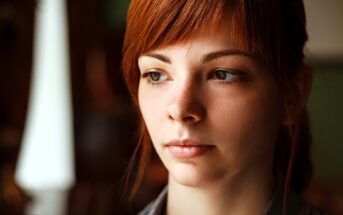 A young woman with red hair and a contemplative expression looks slightly away from the camera, set against a softly blurred background.