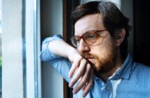 A person with glasses and a beard looks thoughtfully out of a window, leaning their head on one hand. They wear a light blue shirt, and natural light illuminates their face, creating a reflective atmosphere.