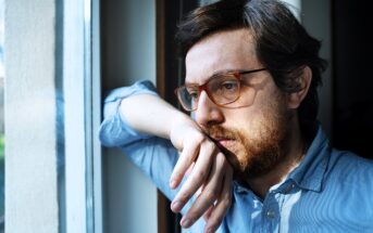 A person with glasses and a beard looks thoughtfully out of a window, leaning their head on one hand. They wear a light blue shirt, and natural light illuminates their face, creating a reflective atmosphere.