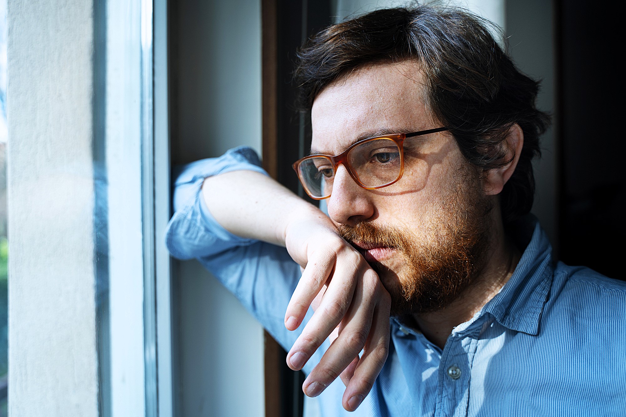 A person with glasses and a beard looks thoughtfully out of a window, leaning their head on one hand. They wear a light blue shirt, and natural light illuminates their face, creating a reflective atmosphere.