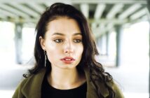 A woman with long, dark hair gazes intently to the side. She is wearing a green jacket and dangling earrings, standing under a structure with beams. The background is softly blurred, emphasizing her thoughtful expression.