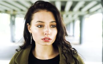 A woman with long, dark hair gazes intently to the side. She is wearing a green jacket and dangling earrings, standing under a structure with beams. The background is softly blurred, emphasizing her thoughtful expression.