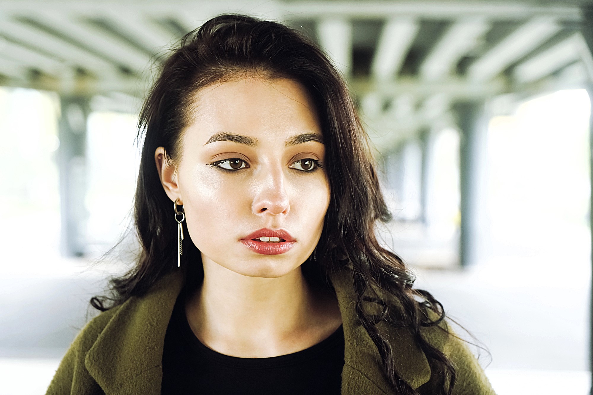 A woman with long, dark hair gazes intently to the side. She is wearing a green jacket and dangling earrings, standing under a structure with beams. The background is softly blurred, emphasizing her thoughtful expression.