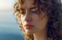 A woman with curly hair gazes contemplatively into the distance. Her expression is thoughtful against a blurred ocean backdrop.