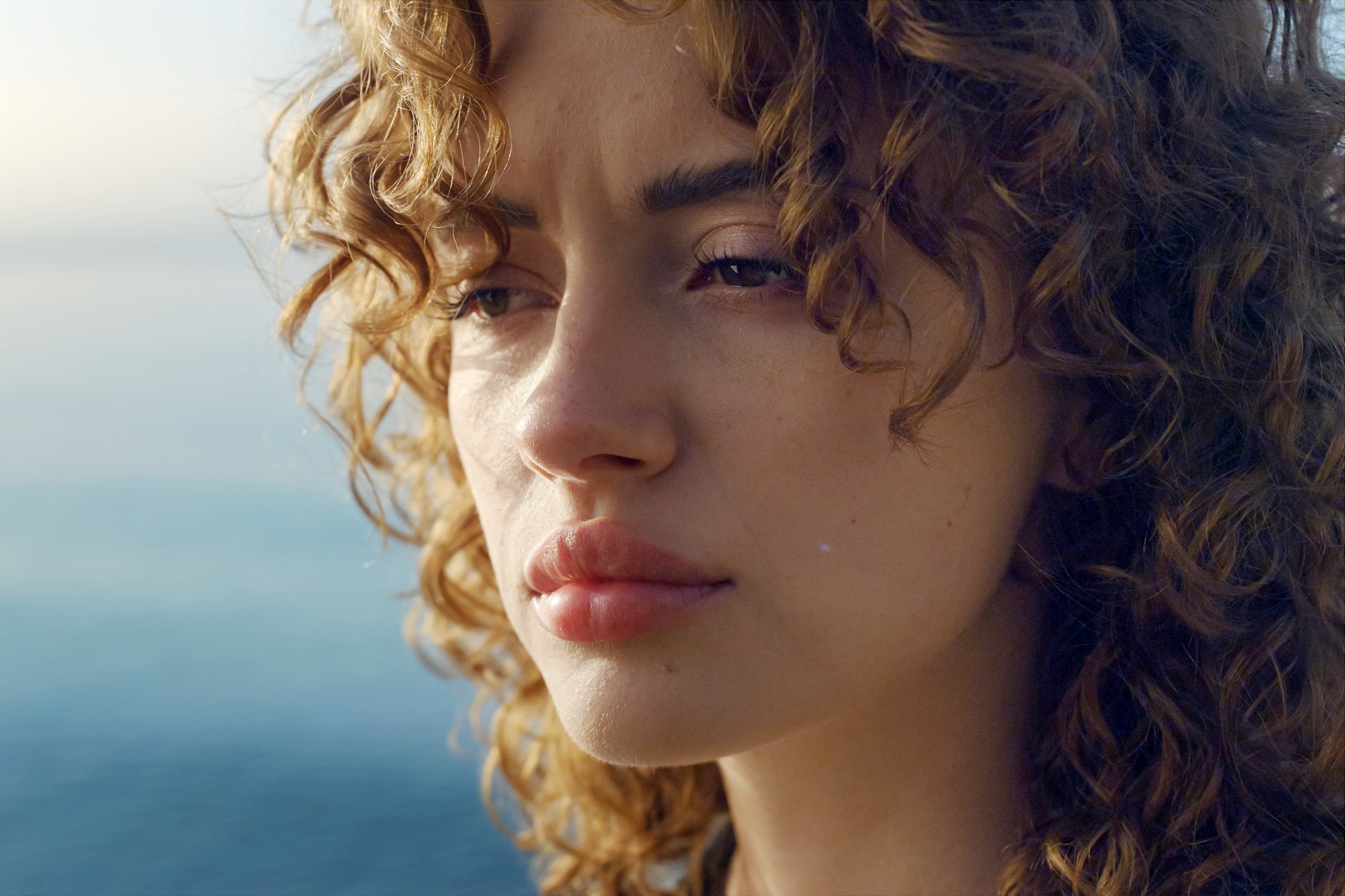 A woman with curly hair gazes contemplatively into the distance. Her expression is thoughtful against a blurred ocean backdrop.