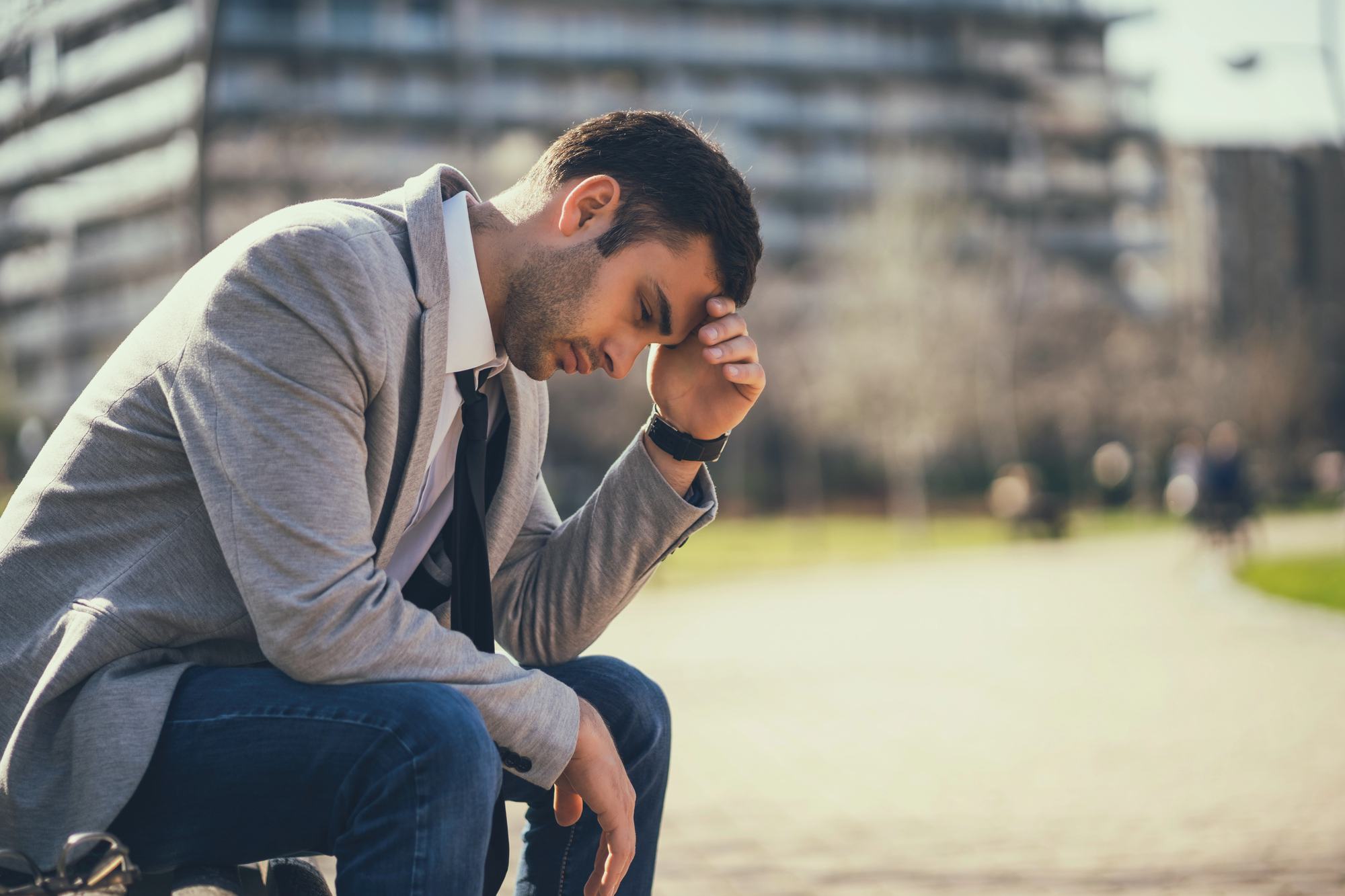 A man in a gray jacket and jeans sits on a bench with his head resting on his hand, appearing thoughtful or stressed. The background shows a blurred urban setting with buildings and trees.