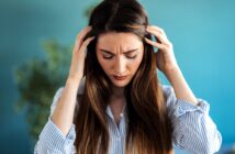 A woman with long brown hair and wearing a striped shirt holds her head with both hands, appearing stressed or upset. A blurred green plant is visible in the background with a turquoise wall.