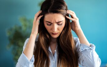 A woman with long brown hair and wearing a striped shirt holds her head with both hands, appearing stressed or upset. A blurred green plant is visible in the background with a turquoise wall.