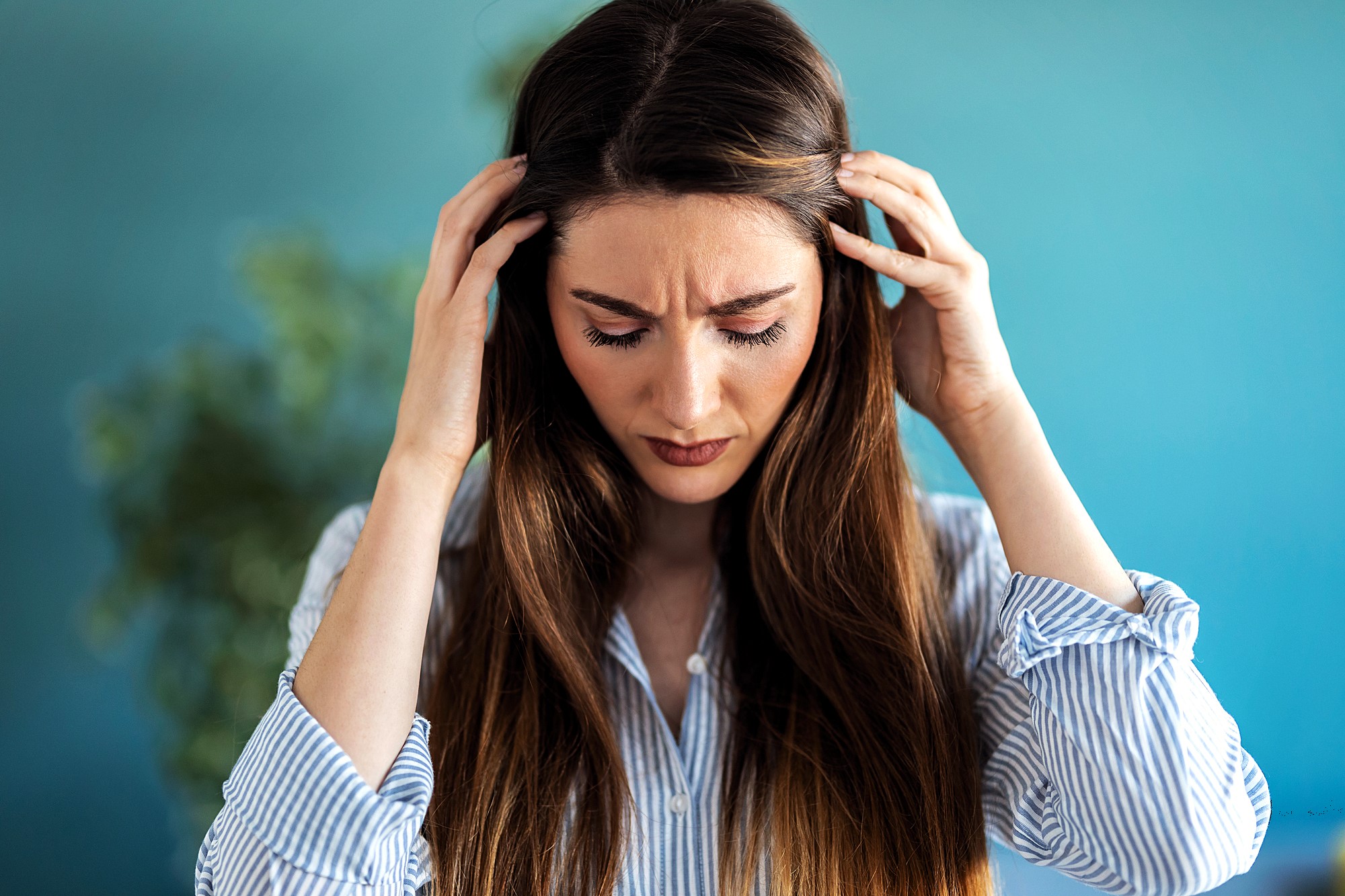 A woman with long brown hair and wearing a striped shirt holds her head with both hands, appearing stressed or upset. A blurred green plant is visible in the background with a turquoise wall.