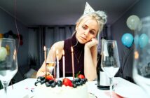 A woman wearing a party hat looks tired and contemplative, resting her chin on her hand. A birthday cake with lit candles and fruit sits in front of her, surrounded by empty wine glasses and a champagne bottle. Balloons decorate the background.