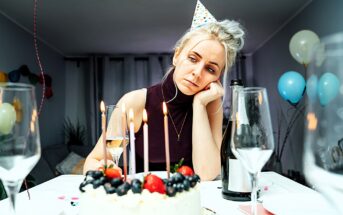 A woman wearing a party hat looks tired and contemplative, resting her chin on her hand. A birthday cake with lit candles and fruit sits in front of her, surrounded by empty wine glasses and a champagne bottle. Balloons decorate the background.