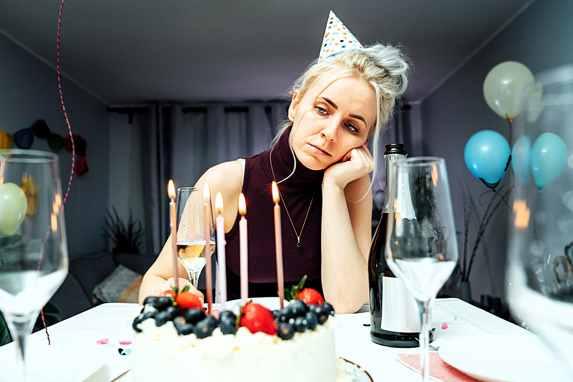 A woman wearing a party hat looks tired and contemplative, resting her chin on her hand. A birthday cake with lit candles and fruit sits in front of her, surrounded by empty wine glasses and a champagne bottle. Balloons decorate the background.