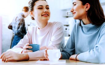 Two women are sitting together at a table, smiling and holding mugs. One woman has a blue mug, and the other has a white cup. They appear to be enjoying a conversation in a bright setting. A person in the background is wearing a plaid shirt.