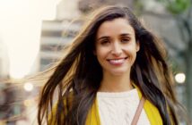 A woman with long brown hair smiles warmly at the camera. She is wearing a yellow jacket over a white sweater. The background shows an urban setting with blurred buildings and trees.