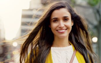 A woman with long brown hair smiles warmly at the camera. She is wearing a yellow jacket over a white sweater. The background shows an urban setting with blurred buildings and trees.