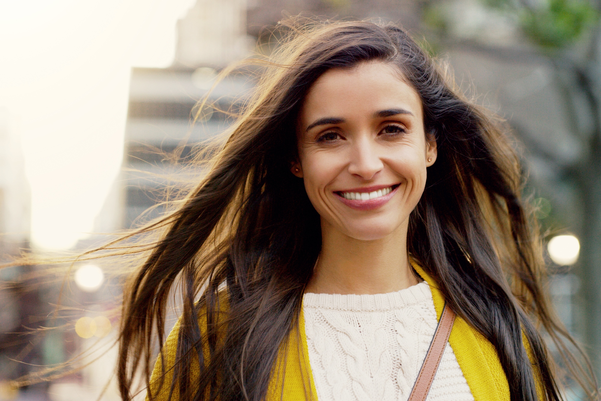 A woman with long brown hair smiles warmly at the camera. She is wearing a yellow jacket over a white sweater. The background shows an urban setting with blurred buildings and trees.