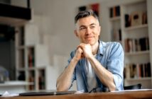 A man with short gray hair is sitting at a table, smiling softly. He rests his chin on his hands, wearing a light blue denim shirt over a white t-shirt. The background features shelves filled with books.