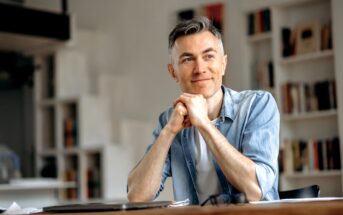 A man with short gray hair is sitting at a table, smiling softly. He rests his chin on his hands, wearing a light blue denim shirt over a white t-shirt. The background features shelves filled with books.