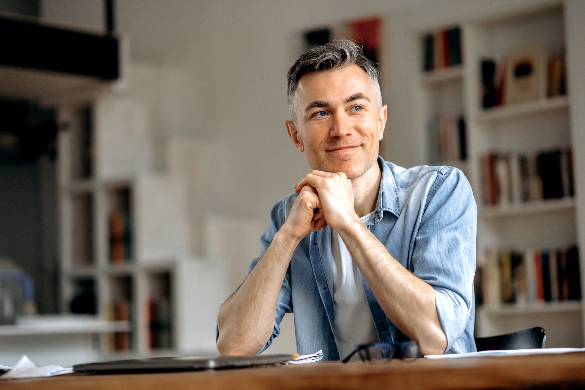 A man with short gray hair is sitting at a table, smiling softly. He rests his chin on his hands, wearing a light blue denim shirt over a white t-shirt. The background features shelves filled with books.