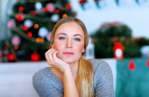 A woman with long blonde hair and a thoughtful expression rests her chin on her hand. She wears a gray sweater. In the background, there's a blurred Christmas tree with red and white ornaments and string lights.