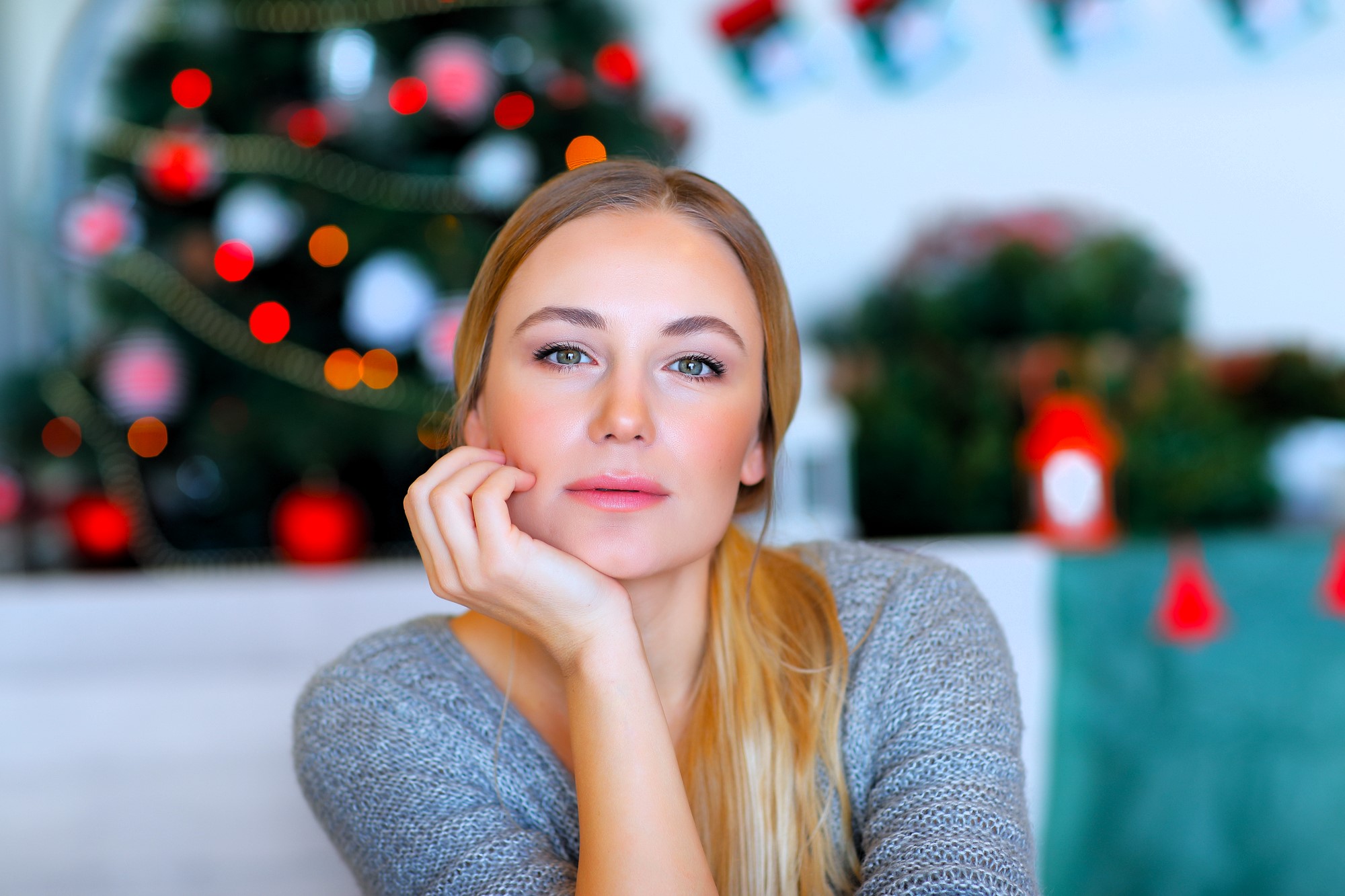 A woman with long blonde hair and a thoughtful expression rests her chin on her hand. She wears a gray sweater. In the background, there's a blurred Christmas tree with red and white ornaments and string lights.