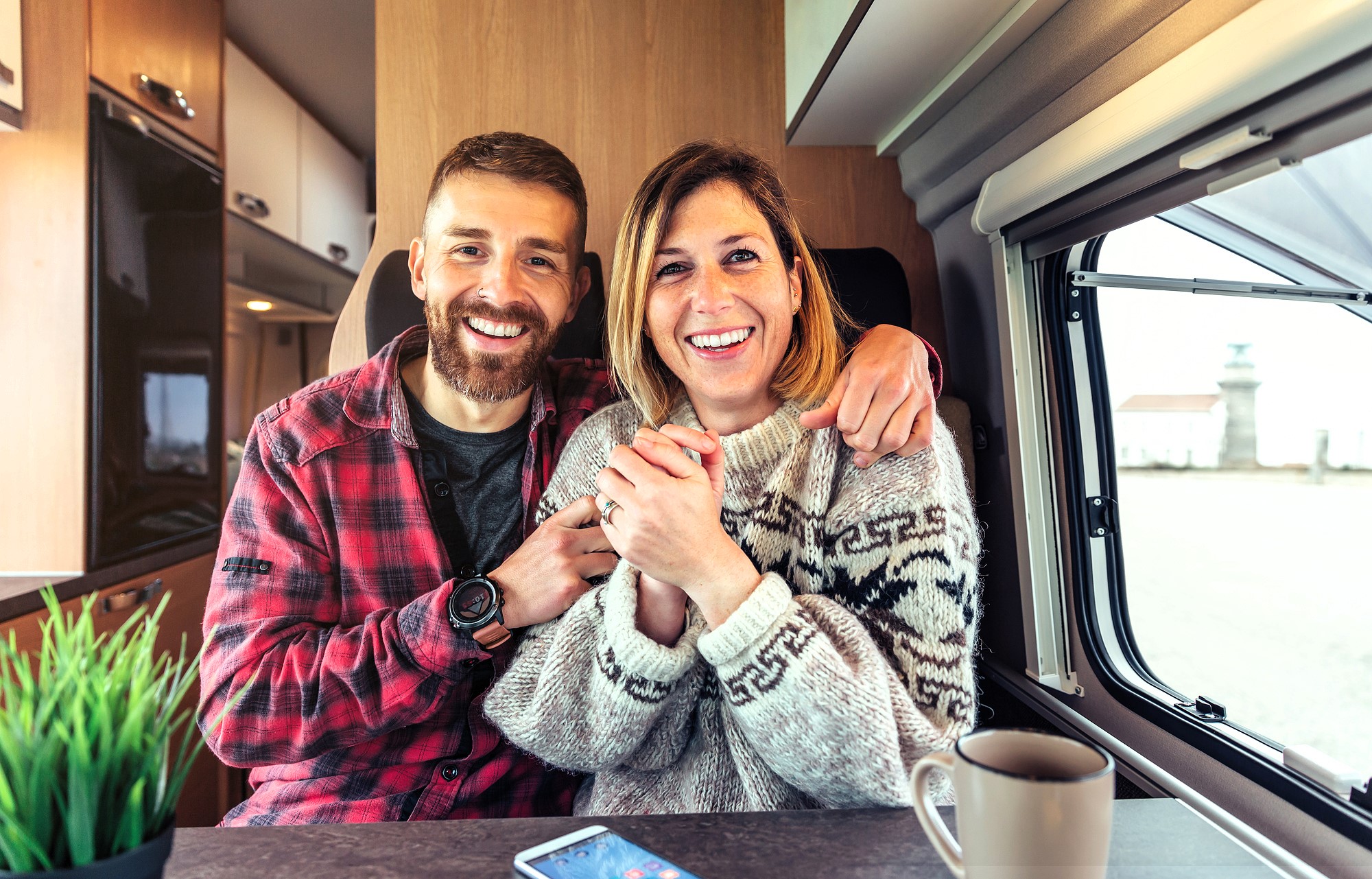 A couple sits inside a cozy camper van, smiling brightly. The man wears a plaid shirt, and the woman wears a patterned sweater. They sit at a table with a cup of coffee and a tablet. Outside the window, a lighthouse is visible. A small plant decorates the table.