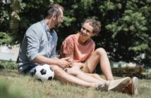 Two people sit on the grass with a soccer ball beside them, smiling and talking. One person wears a light blue shirt and jeans, the other wears a peach shirt and shorts. They appear relaxed, enjoying a sunny day outdoors with trees in the background.