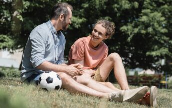 Two people sit on the grass with a soccer ball beside them, smiling and talking. One person wears a light blue shirt and jeans, the other wears a peach shirt and shorts. They appear relaxed, enjoying a sunny day outdoors with trees in the background.