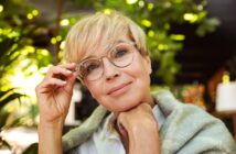 A person with short blonde hair and glasses smiles gently while resting their chin on their hand. They are wrapped in a shawl, with a background of lush greenery and soft, filtered light.