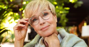 A person with short blonde hair and glasses smiles gently while resting their chin on their hand. They are wrapped in a shawl, with a background of lush greenery and soft, filtered light.