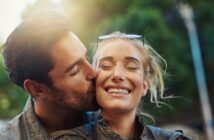 A man kisses a smiling woman on the cheek outdoors. Both are dressed casually, and the woman has sunglasses on her head. The background features blurred greenery and sunlight.