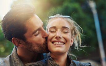 A man kisses a smiling woman on the cheek outdoors. Both are dressed casually, and the woman has sunglasses on her head. The background features blurred greenery and sunlight.