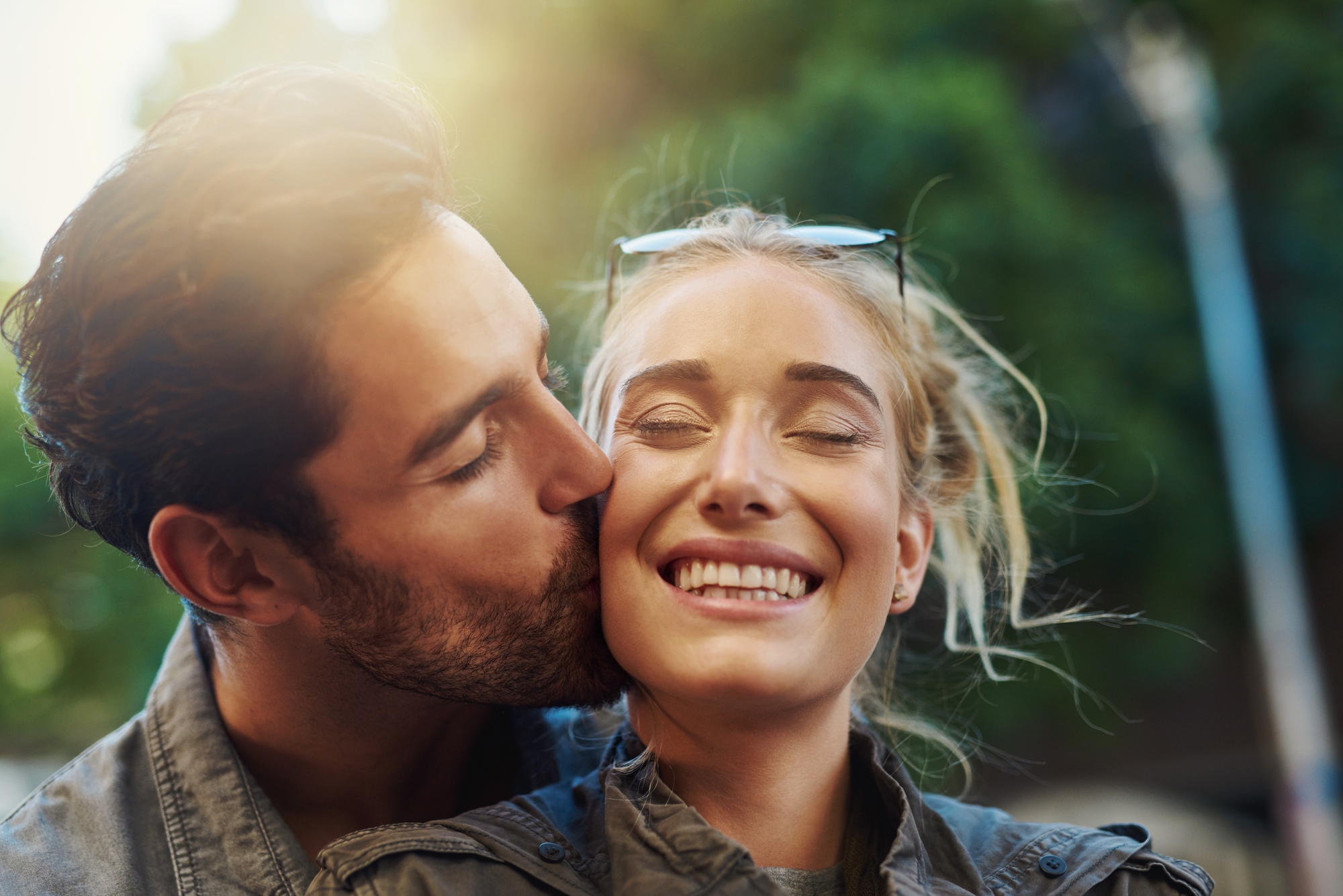 A man kisses a smiling woman on the cheek outdoors. Both are dressed casually, and the woman has sunglasses on her head. The background features blurred greenery and sunlight.