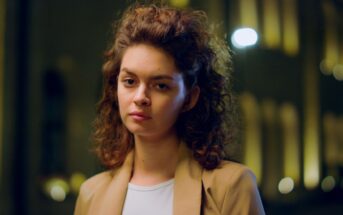 A person with curly hair wearing a tan coat over a white shirt is standing outdoors at night. The background is softly blurred with warm yellow lights.