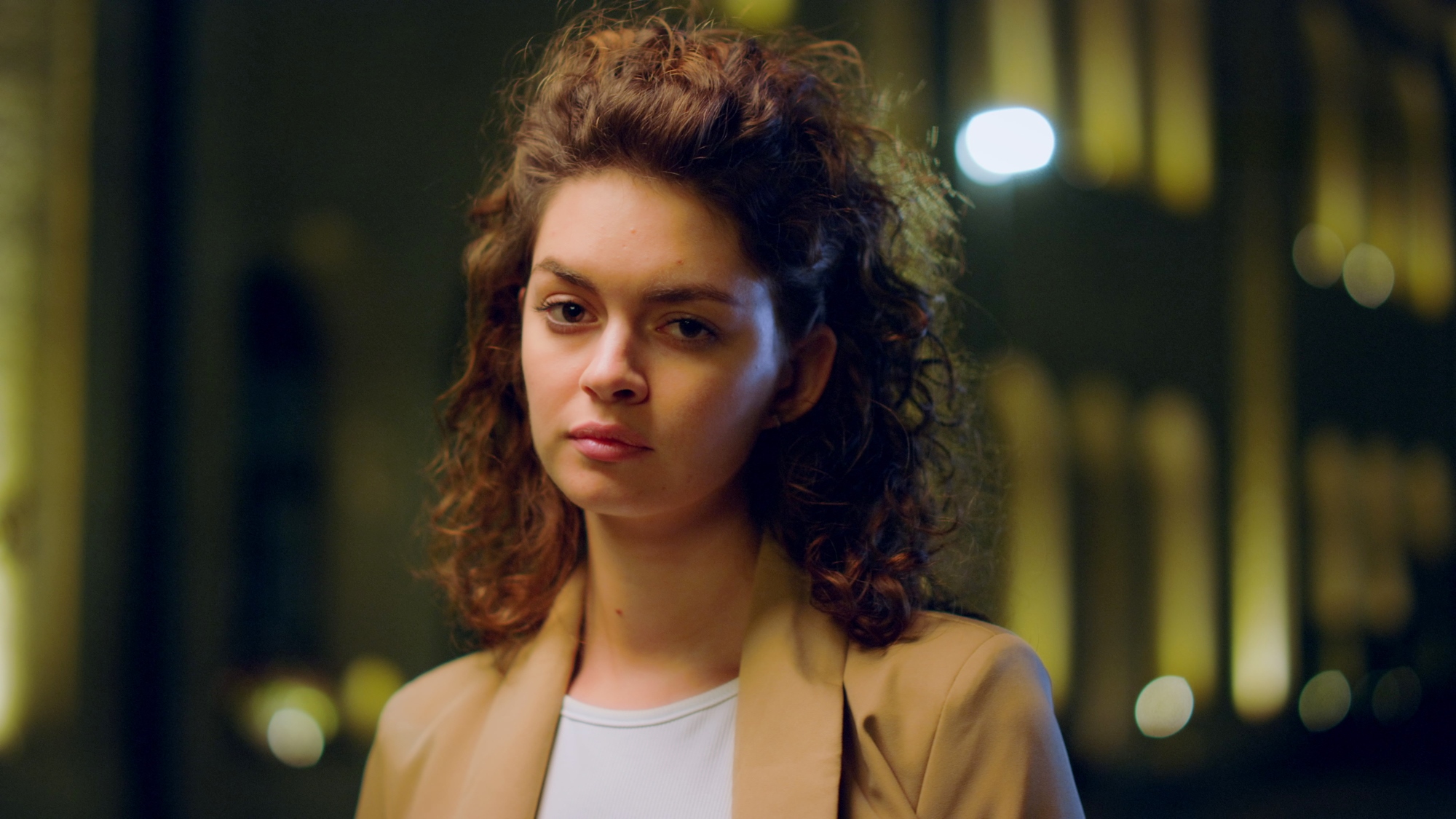 A person with curly hair wearing a tan coat over a white shirt is standing outdoors at night. The background is softly blurred with warm yellow lights.