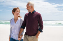 A couple walks hand in hand along a sunny beach. The woman has red hair and wears a light cardigan over a white blouse, while the man has gray hair and wears a maroon sweater over a dark shirt. They are smiling at each other.