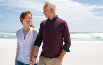 A couple walks hand in hand along a sunny beach. The woman has red hair and wears a light cardigan over a white blouse, while the man has gray hair and wears a maroon sweater over a dark shirt. They are smiling at each other.