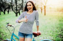 A woman in a striped shirt and denim shorts stands next to a bicycle in a grassy park, smiling and giving a thumbs-up. The background features trees and sunlight filtering through the branches.